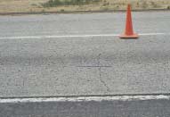 FIGURE 98.  Distress Type CRCP 3 - Low Severity Transverse Cracking, Color photograph of continuously reinforced concrete pavement with distress type CRCP 3 - low severity transverse cracking.  Two lanes of pavement are depicted, and the lane in the foreground has a transverse crack  from the inner wheel path to the lane edge.  The crack is approximately 12 mm wide, as indicated by the 500-mm scale pictured in the center of the photograph, with no evident spalling.