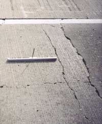 FIGURE 125.  Distress Type CRCP 12 - Moderate Severity Punchout, Color photograph of continuously reinforced concrete pavement with distress type CRCP 12 - moderate severity punchout.  A single punchout is pictured, in which 2 parallel transverse cracks have been intersected by a longitudinal crack to form the rectangular area next to the pavement joint.  Areas with spalling up to approximately 75 mm and faulting up to approximately 12 mm are evident, as indicated by the 500-mm scale in the center of the photograph.