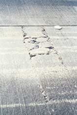 FIGURE 126.  Distress Type CRCP 12 - High Severity Punchout, Color photograph of continuously reinforced concrete pavement with distress type CRCP 12 - high severity punchout.  An area of punchouts is pictured, in which 2 parallel transverse cracks have been intersected by several longitudinal cracks to form rectangular areas next to the pavement joint.  There is significant spalling and loss of concrete, and some pieces of the surface are loose. 