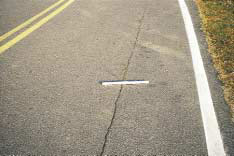 FIGURE 14.  Distress Type ACP 4a - Moderate Severity Longitudinal Cracking in the Wheel Path, Color photograph of asphalt concrete pavement with distress type ACP 4a - moderate severity longitudinal cracking in the wheel path.  As indicated by the 500-mm scale pictured in the center of the photograph, the crack is approximately 18 mm wide, and extends through the outer wheel path along the entire length of the lane.
