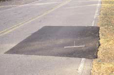 FIGURE 24.  Distress Type ACP 7 - Low Severity Patch, Color photograph of asphalt concrete pavement with distress type ACP 7 - low severity patch.  The large rectangular patch extends across one lane of a two-lane highway from the edge of the road to the middle of the inner wheel path and shows no visible deterioration.  The width of the patch is approximately 2 m and the length is approximately 2 m, as indicated by the 500-mm scale near the center of the photograph. 