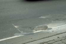 FIGURE 25.  Distress Type ACP 7 - High Severity Patch, Color photograph of asphalt concrete pavement with distress type ACP 7 - high severity patch.   The patched area is located near the shoulder and contains two large patches that are generally round in shape and appear to be potholes that were repaired. There is extensive cracking and deterioration within and surrounding the patches.
