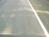 FIGURE 46.  Distress Type ACP 15 - Fine Material Left on Surface by Water Bleeding and Pumping, Color photograph of asphalt concrete pavement with distress type ACP 15 - fine material left on surface by water bleeding and pumping.  The photo shows a lane of highway with two transverse cracks that have been stained by water and fine material that has seeped through the cracks from beneath the pavement.