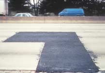 FIGURE 86.  Distress Type JCP 15 - Large, Low Severity Asphalt Concrete Patch, Color photograph of jointed portland cement concrete pavement with distress type JCP 15 - large, low severity asphalt concrete patch.  The L-shaped patch extends across two lanes, and low severity deterioration of the patch, including cracking and loss of materials, is visible at the edges of the patch.