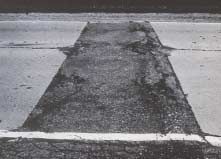 FIGURE 87.  Distress Type JCP 15 - Large, Low Severity Asphalt Concrete Patch, Color photograph of jointed portland cement concrete pavement with distress type JCP 15 - large, low severity asphalt concrete patch.  The rectangular patch extends across two lanes and across the edge stripe to the shoulder.   High severity deterioration of the patch, including cracking, spalling, scaling and loss of materials, is visible across large sections of the patch.