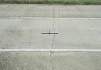 FIGURE 99.  Distress Type CRCP 3 - Moderate Severity Transverse Cracking, Color photograph of continuously reinforced concrete pavement with distress type CRCP 3 - moderate severity transverse cracking. Three lanes of pavement are depicted, and there is a continuous transverse crack that extends across the width of the two lanes in the foreground.  The crack is approximately 25 mm wide, as indicated by the 500-mm scale pictured in the center of the photograph, with spalling along approximately 25% of the crack length.