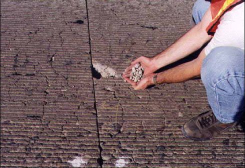 The photograph shows a concrete pavement at a transverse joint with numerous deep cracks and an actual hole in the pavement caused by cracking. An individual is shown in the photograph holding pieces of he disintegrated pavement from the hole.