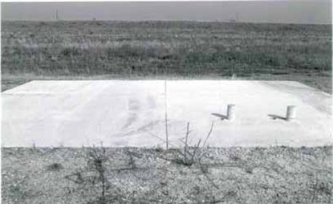 Test panels for mixture M N 7 showing cores taken in 1994. The figure is a faded black-and-white photograph of four 2.3 by 3.0 meter concrete slabs with minor distress. The photo was taken at a 45-degree angle. Two cylinder blocks sit on the lower right slab. There is some minor distress on the surface that might be caused by construction. 