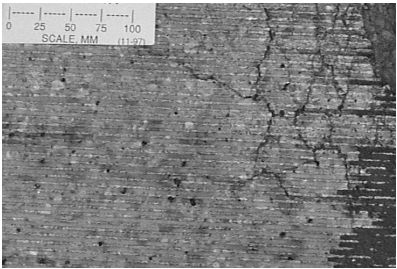 This black-and-white photograph shows horizontal grooves running on a parallel pattern across the surface. A large alligator crack begins on the right side of the concrete slab. Large sand particles are resting on the center. On the top left corner is a piece of paper with a measuring scale in millimeters. This surface map cracking was noted in 1997.