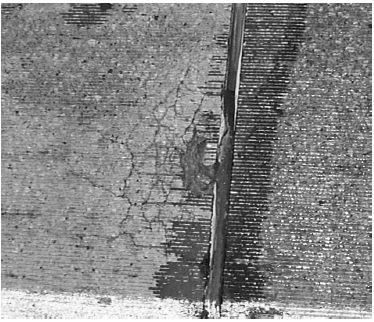 This figure is a black-and-white photograph of a concrete slab. On the bottom of the picture is a white, horizontal paint strip. There is a long vertical crack in the center. The left side of the vertical crack extends into alligator cracks. Brooming is visible. There are horizontal grooves lining the surface. The cracking was generally worse in the wheelpath near the transverse joints between the patches and the original pavement.