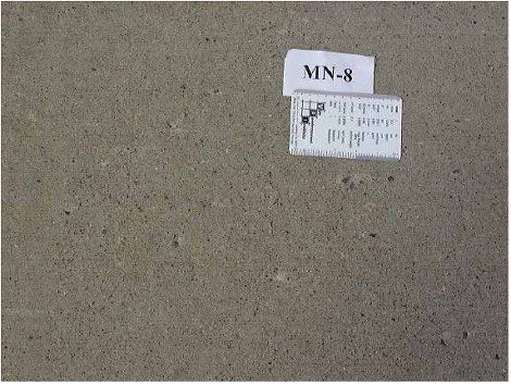 This figure is a close-up color photograph of one concrete slab. The slab has a measuring scale lying on top of it. There are a few small popouts scattered throughout the slab.