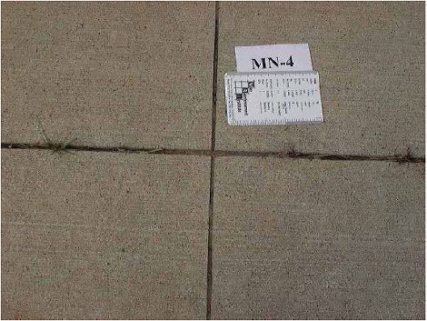 This figure is a color photograph of four concrete slabs. There are specks all over the surface, probably small popouts. There is a small ruler lying on the top right slab. There are no significant spalling or scaling present. The surface is in overall good condition.