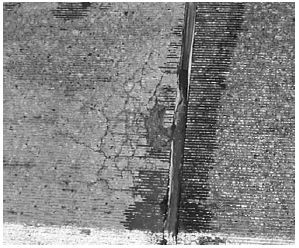 This figure is a black-and-white photograph of cracking in concrete. The concrete has horizontal grooV E S along the entire surface. On the center of the surface is a wide vertical split. To the left of the split are alligator cracks. Some of the surface has come off on the alligator cracks