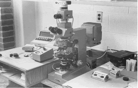 The microscope is positioned in the center of the photo. In the right foreground are two pushbutton counters. The camera and exposure meter are on top of the microscope. The control for the exposure meter is between the microscope and the automatic point counter keyboard in the left background. 