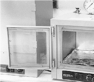 A square oven with a transparent glass front door is shown with the door open. Two sample trays, each containing a thin-section, are in the oven and ready to be vacuum-oven dried
