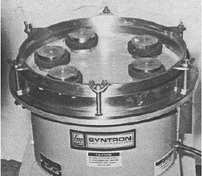 Photo shows a cylindrical vibratory machine that rests vertically on a countertop and is firmly positioned by a wider metal base. A lapping pad is mounted on the vibrating surface of the machine. Over the lapping pad are six individual circular metal weights, each holding a thin section in place for polishing. The weights are free to move over the surface of the lapping pad and have sponge rubber bumper rings mounted on them for shock absorption. A railing around the lapping surface keeps the weights and their samples from slipping off the surface of the vibrating pad. Reference is made to Walker and Marshall, 1979.