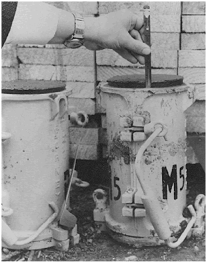 After it was cast in the cylinder mold, the concrete increased in volume as hydrogen gas evolved from the chemical reaction of aluminum (from an aluminum delivery pipe) with the alkaline fluids of the fresh cement paste. The photo shows two 150 by 300 millimeter heavy steel cylinder molds with concrete rising above the top rim of the molds. To give scale a hand is shown holding a pen on the top rim of one of the molds to show that the concrete has risen about 10 millimeters.
