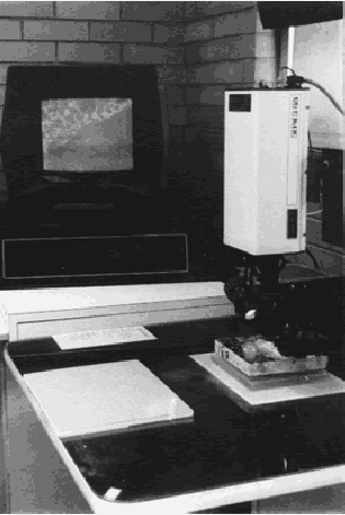 Photo shows an assembly of video camera with magnifying lens mounted vertically and scanning a concrete slice.  A computer screen in the background shows the progress of the analysis. (Photograph by R.H. Howe, courtesy of the Pennsylvania Department of Transportation.)