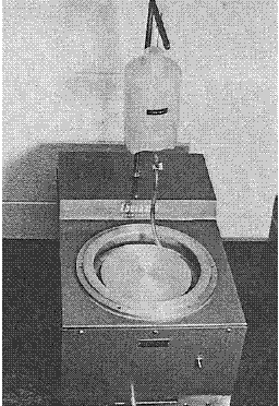 A turntable with a 200 millimeter disk used for rough grinding rock and concrete specimens. A jar for water or another lubricant is suspended approximately 0.3 meter above the disk and is used for dripping water or other lubricants during grinding. Grinding compounds may be added manually with plastic dispensers.
