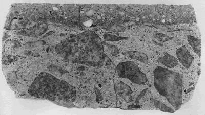 The slice of concrete shown is a cross section of concrete bridge deck with a stratum of latex-modified concrete overlay placed above a substrate of normal concrete. Cracking has formed, not exactly at the strata boundary but slightly below it. Also, a vertical crack in the substrate continues through the latex concrete and up to the wearing surface. The specimen is 100 millimeters in width.