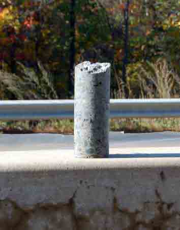 Photo. Close-up of extracted core from a concrete barrier. This photo shows a concrete core that was extracted from the barrier; the core is 10 centimeters (4 inches) in diameter and about 30 centimeters (12 inches) in length. The core sits on top of a concrete barrier.