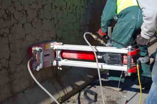 Photo. Coring equipment being used to extract a (10-centimeter) 4-inch diameter core from a concrete barrier. This photo shows a person using horizontal coring equipment to extract a concrete core from an ASR-affected concrete barrier. 