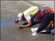 Figure 13. Photo. Expansion Measurements Being Conducted After Pavement Was Treated With Lithium Nitrate. This photo shows two members of the research team taking expansion measurements on the surface of an ASR-affected pavement recently treated with lithium. One member is taking measurements from the surface pavement using a measurement gage, and the other member is recording the data. The pavement surface is wet.