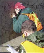 Figure 14. Photo. Cracking Measurements Performed on a Concrete Barrier near Leominster, MA. This photo shows two members of the research team taking crack mapping data from an ASR-affected concrete barrier. One member is viewing the barrier through a handheld microscopic device; the other member is noting the measurements.