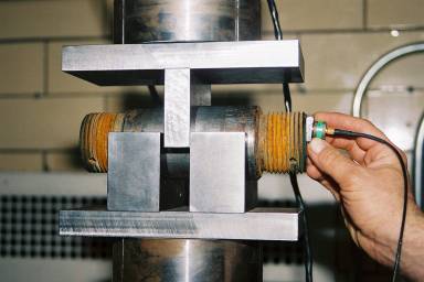 Photograph of pulse-echo setup using 14-degree transducer.  Color Photograph.  This figure is a color photograph of the pulse-echo setup with a 14-degree transducer used during the acoustic coupling tests.  The photograph shows a hand holding the transducer at the end of the pin, which is held in place by a test frame.