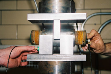 Photograph of pitch-catch setup using 0-degree transducers.  Color Photograph.  This figure is a color photograph of a pitch-catch setup with two 0-degree transducers used during the acoustic coupling tests.  The photograph shows a hand holding the transmitting transducer at the right end of the pin, which is held in place by a test frame.  Another hand is holding the receiving transducer at the left side of the test frame.