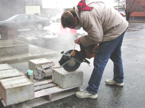 Figure 27. Cutting a test slab with a gas-powered saw. Photo. This picture shows a man outdoors working with five slabs on a wooden pallet. He is wearing a protective mask and is cutting a test slab through the top with a gas-powered rotary saw.