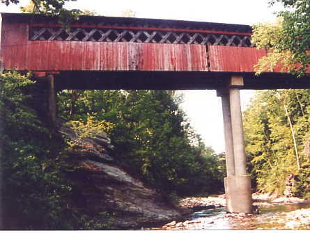 This picture taken from ground level looking up shows part of a long span Town lattice covered bridge, painted red, set on a tall concrete pier near midspan. The pier actually supports 3 steel beams that have been added.