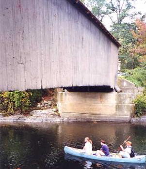 This closeup partial view of a covered bridge shows three people in a canoe about to pass under the span. The bridge sits on concrete abutments set close to the water with narrow clearance.