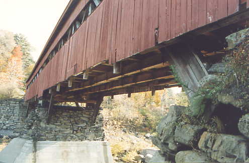 This closeup of the underside of a Town lattice truss covered bridge shows the typical components: stone abutment shored up underneath with concrete walls, truss diagonals, bottom chord with vertical tails and stringers, and siding painted red.