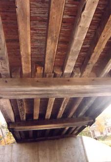 The picture shows the underside of a bridge set on a concrete abutment. The floor beams are horizontal and heavy and are well-spaced while the stringers are vertical, and support the bridge decking.