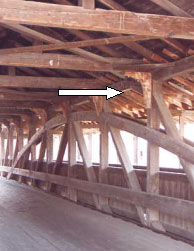 The white arrow points to a short wood diagonal made from the juncture of a tree root and trunk, modeled after those used in old ships. They are located in the upper corner of the bridge that connects the tie beam to the web members of the truss. These knee braces occur at regular intervals to brace and keep the bridge cross section square along its length. These members are the most vulnerable to traffic damage but this configuration presents less intrusion into the lane.