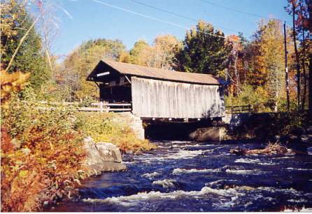 This picture shows a side view of a covered bridge with a protruding portal and approach rails over a white water river running high, and the autumn colors still visible.