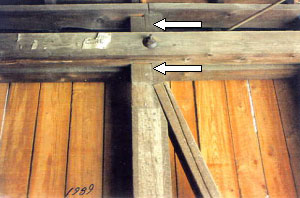 The truss diagonals bear on shoulders cut into the sides of the vertical tension members that are subject to overstressed shear forces along the grain. The picture shows two white arrows pointing to the vertical shift of the right half of the post where separation and slippage shows at the notch for the diagonal.