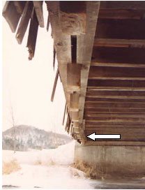 The underside longitudinal view of this bridge shows an arrow pointing to the bowing of the inside of the same chord after the tail has broken off. The chord has been pushed out from under the floor beams at midspan. Only the longitudinal decking spiked to the floor beams holds the floor in place.
