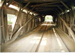 This picture shows the interior of a Burr arch bridge with hardwood running planks following the bridge axis, positioned along wheel paths in two different runs, 0.91 meters (3 feet) wide, made of multiple planks for each unit. These members are sacrificed to wear and replaced without having to replace the entire deck. Twin pairs of running planks encourage drivers to slow down to enforce speed restrictions on one-lane bridges.