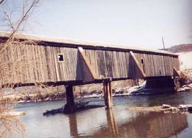 The picture shows a side view of a bridge with piers added after the original construction. These supplemental pile bents are timber telephone poles in a cluster of three driven beneath both trusses with a cap or horizontal member connecting the tops of the piles. Such retrofits are considered temporary because they change truss behavior and not acceptable as neither a historic or esthetic, long-term solution.