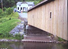 This bridge retains its original stone abutments, but has sheet steel piling around three sides of the masonry to protect against scour. A new concrete cap is visible just below the timber of the bridge. Also along the bank large stone protection protects the slopes from scour.