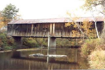 This picture shows an almost full side view of a covered bridge with a concrete pier. The bottom of the siding looks discolored. It is not clear whether there was a pier on the original bridge.
