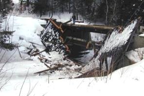 The picture shows the effect of snow without a vehicle load. The bridge is collapsed and fallen on its side with snow surrounding it. The roof is also collapsed and broken in multiple pieces. The text mentions that while uniform snow load is important to consider, often unsymmetrical snow load from drifting causes more problems.
