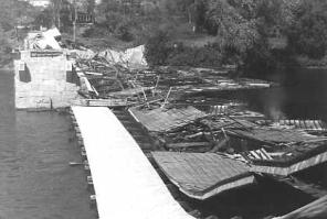 The picture shows a river with the bridge abutment and the bridge deck the only part remaining. The sides and roof are completely destroyed with pieces floating in the river. This collapse occurred after a rehabilitation that extended the siding closer to the eaves of the bridge to decrease ventilation and filled concrete pockets that butted the arches against a flat surface that reduced lateral support.