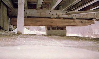 The picture shows the underside of a bridge. A large timber bolster beam parallel to the bottom chord sits on bearing blocks to help provide extra support to the truss beyond the face of the abutment.