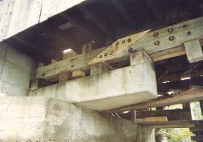 In this underside view of a bridge, the bolster beam is made of concrete and sits on the bridge abutment. The bottom chord shows many bolts with the posts passing through. The wedges are the members that sit directly on top of the concrete bolster.