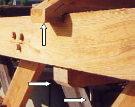 The picture shows two white arrows pointing to transverse tie beams that connect to top chords. The underside of the tie beam that straddles the top chord is notched. The third arrow points to a vertical bolt that goes through the top chord and tie beam. These beams hold the spacing between trusses, help prevent buckling of the top compression chords and maintain the bridge alignment.