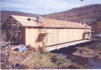 The picture shows a side view of a bridge being rehabilitated. The decking is extended so staging can be set on it. Staging and access ladders are at the entrance and along the bridge length. Two workers are re-roofing and another is cutting boards at the entrance.