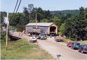 The picture shows a bridge on wooden blocks elevated above the road surface next to a conventional highway bridge. Cars and trucks are parked on the nearby roadway and passersby are observing the bridge under repair.
